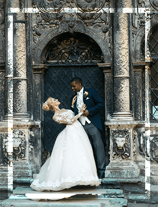 Bride in a white gown and groom in a formal suit embrace playfully at an ornate stone arch, capturing a romantic moment in a historic setting.