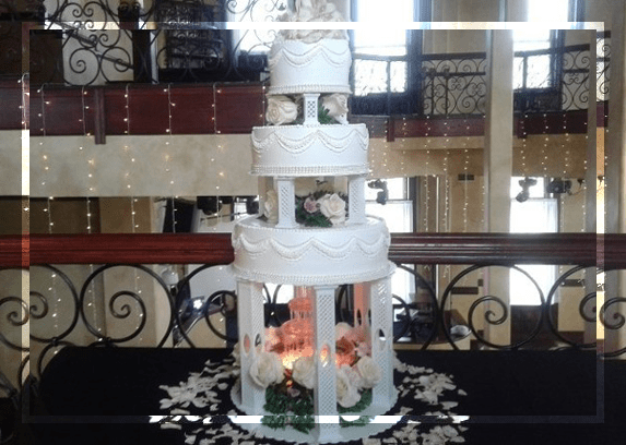 A four-tiered white wedding cake, adorned with delicate roses and greenery, is elegantly displayed on a table, surrounded by soft lighting and decorative accents.