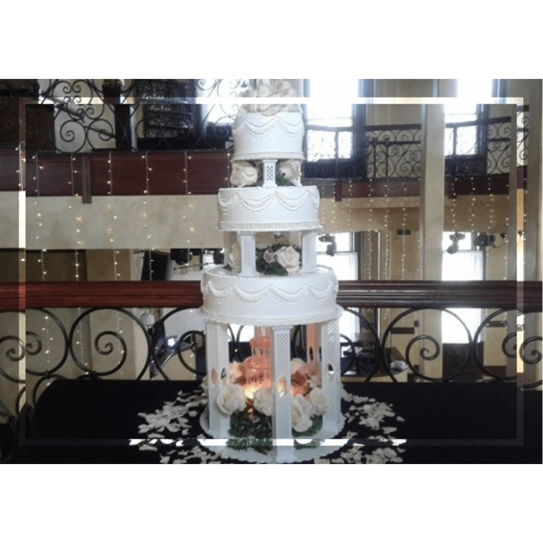 Elegant three-tiered wedding cake with white frosting and decorative roses, displayed in a festive venue with twinkling lights.